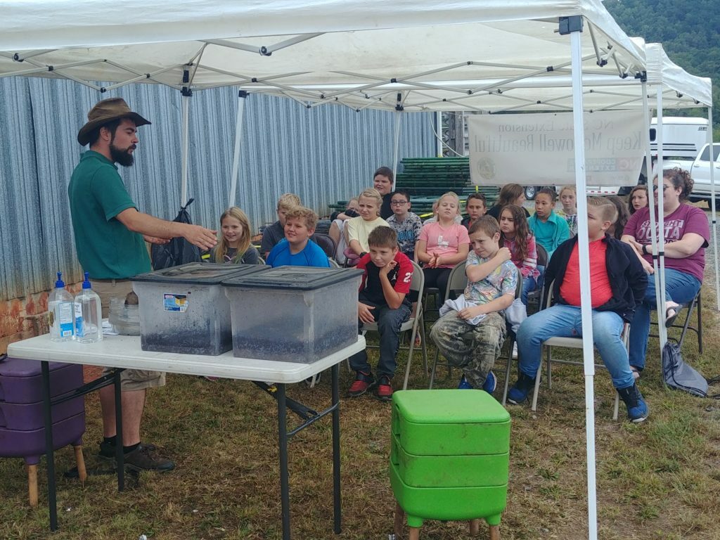 Outdoor compost demo. Instructor shows compost bins while students look on from chairs under a tent. 