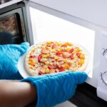 pizza going into microwave with blue potholders on hands